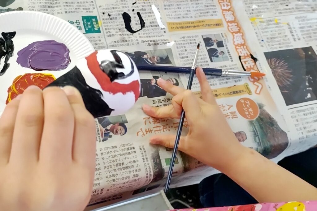 新聞紙の上で、おきあがり小法師に絵付けをする子どもの手